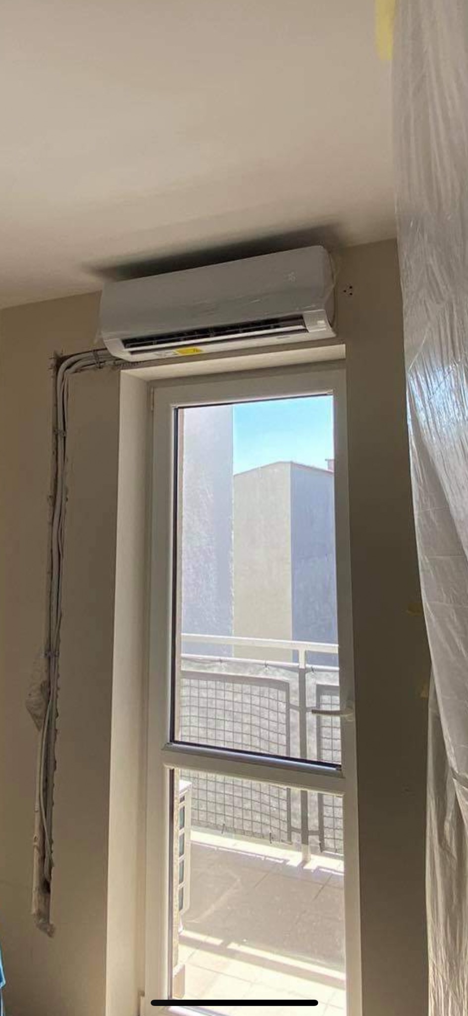 Biały klimatyzator typu split zamontowany nad oknem balkonowym w jasnym pomieszczeniu, widoczne przewody elektryczne w ścianie przed zakryciem, okno zabezpieczone folią malarską.
