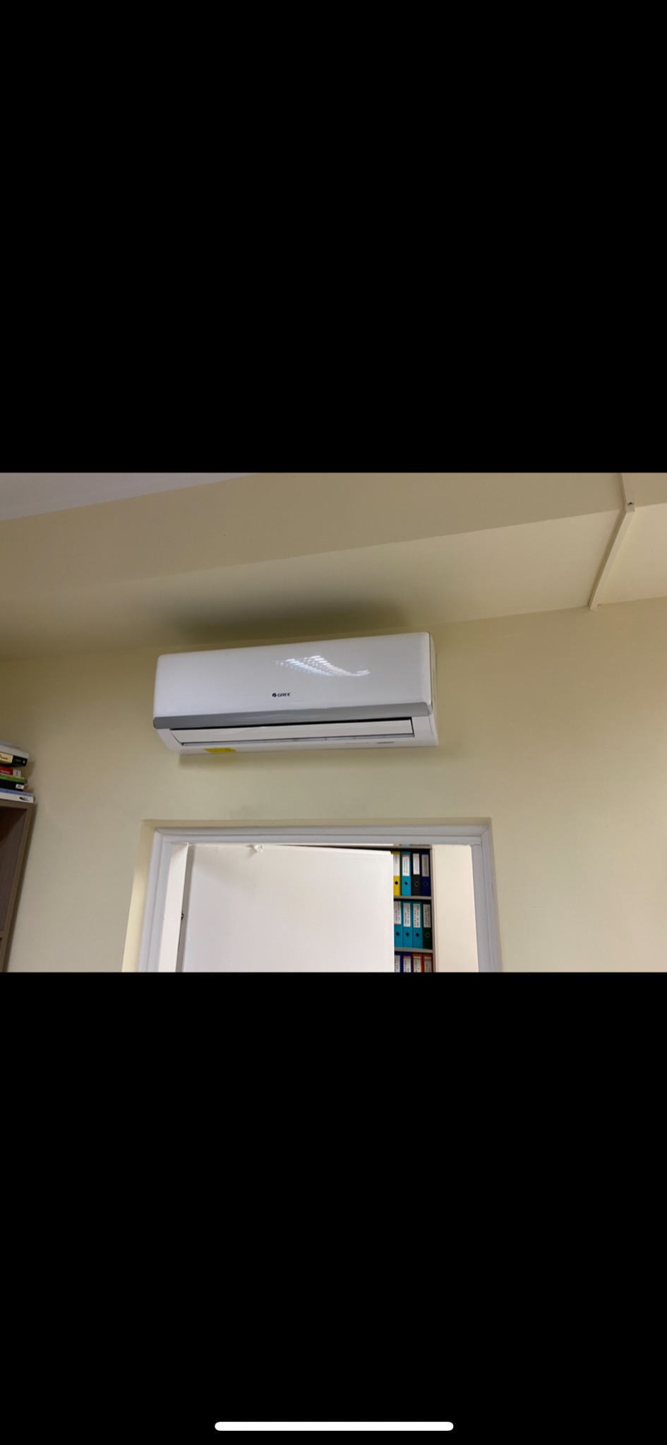 Biały klimatyzator marki Gree zamontowany na beżowej ścianie nad otwartymi drzwiami, widoczne segregatory w tle.