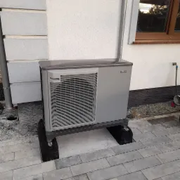 Szara pompa ciepła Nibe F2040 zamontowana na czarnych podstawach antywibracyjnych na tle białej ściany domu z widocznym oknem.