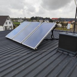 HYDROKAM INSTALACJE W TWOIM DOMU - Trzy panele słoneczne zamontowane na szarym, blaszany dachu domu jednorodzinnego, widok na okoliczne budynki w pochmurny dzień.