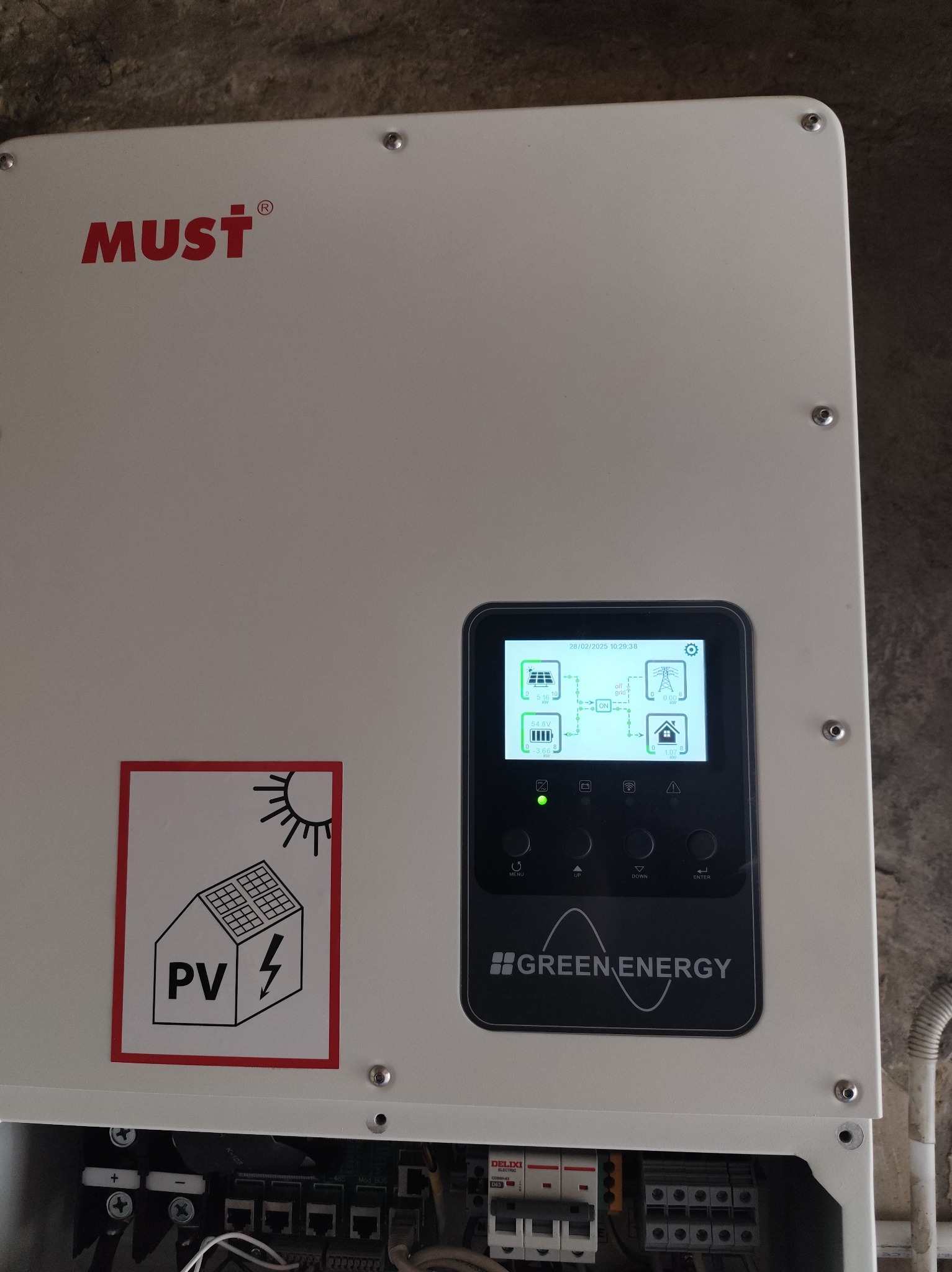 Biała obudowa falownika fotowoltaicznego marki MUST z wyświetlaczem pokazującym przepływ energii, naklejką ostrzegawczą PV i widocznymi elementami instalacji elektrycznej w dolnej części.