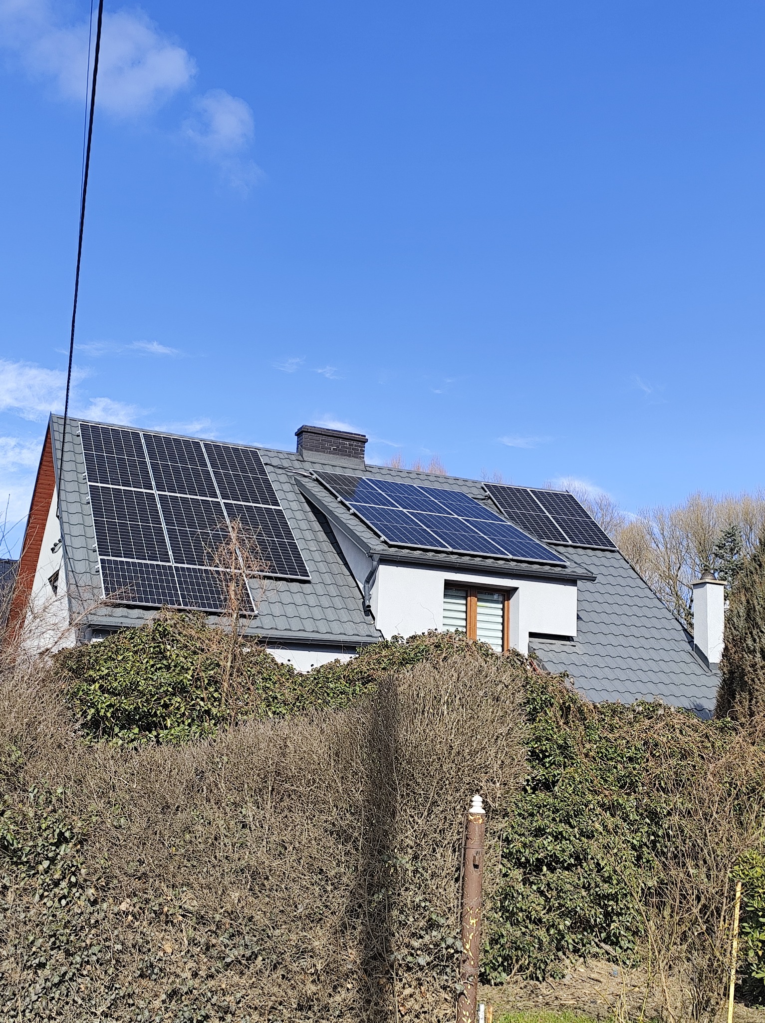 Dom jednorodzinny z panelami słonecznymi zamontowanymi na szarym dachu, częściowo zasłonięty przez gęsty żywopłot. Błękitne niebo w tle.