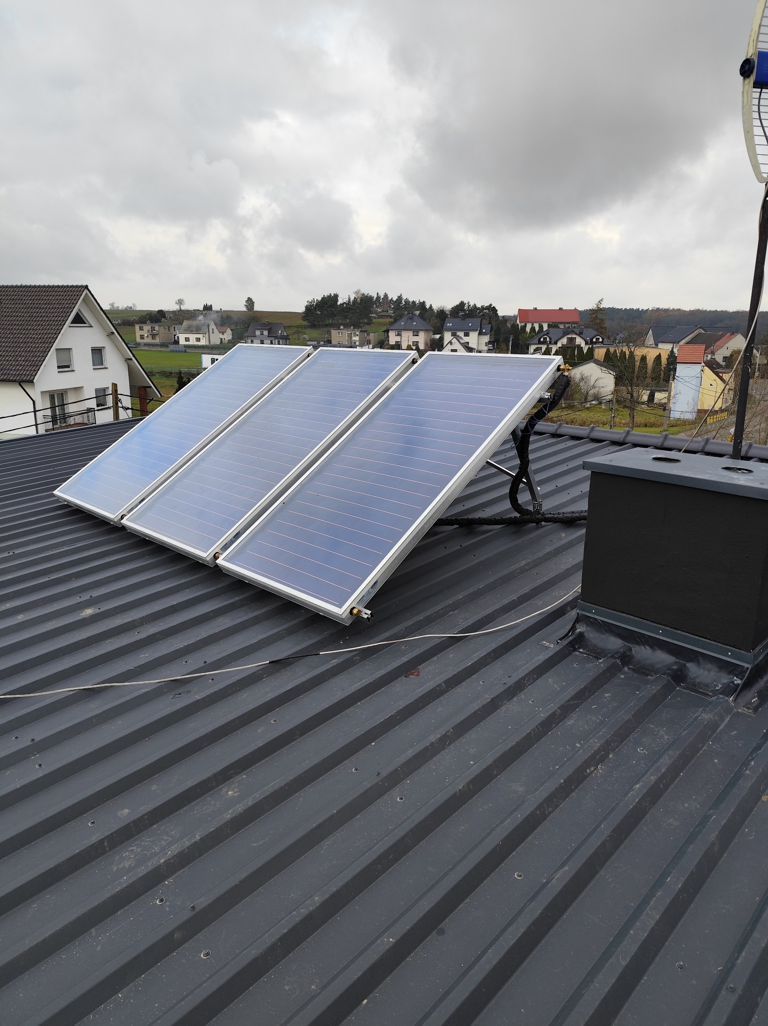 Trzy panele słoneczne zamontowane na szarym, blaszany dachu domu jednorodzinnego, widok na okoliczne budynki w pochmurny dzień.