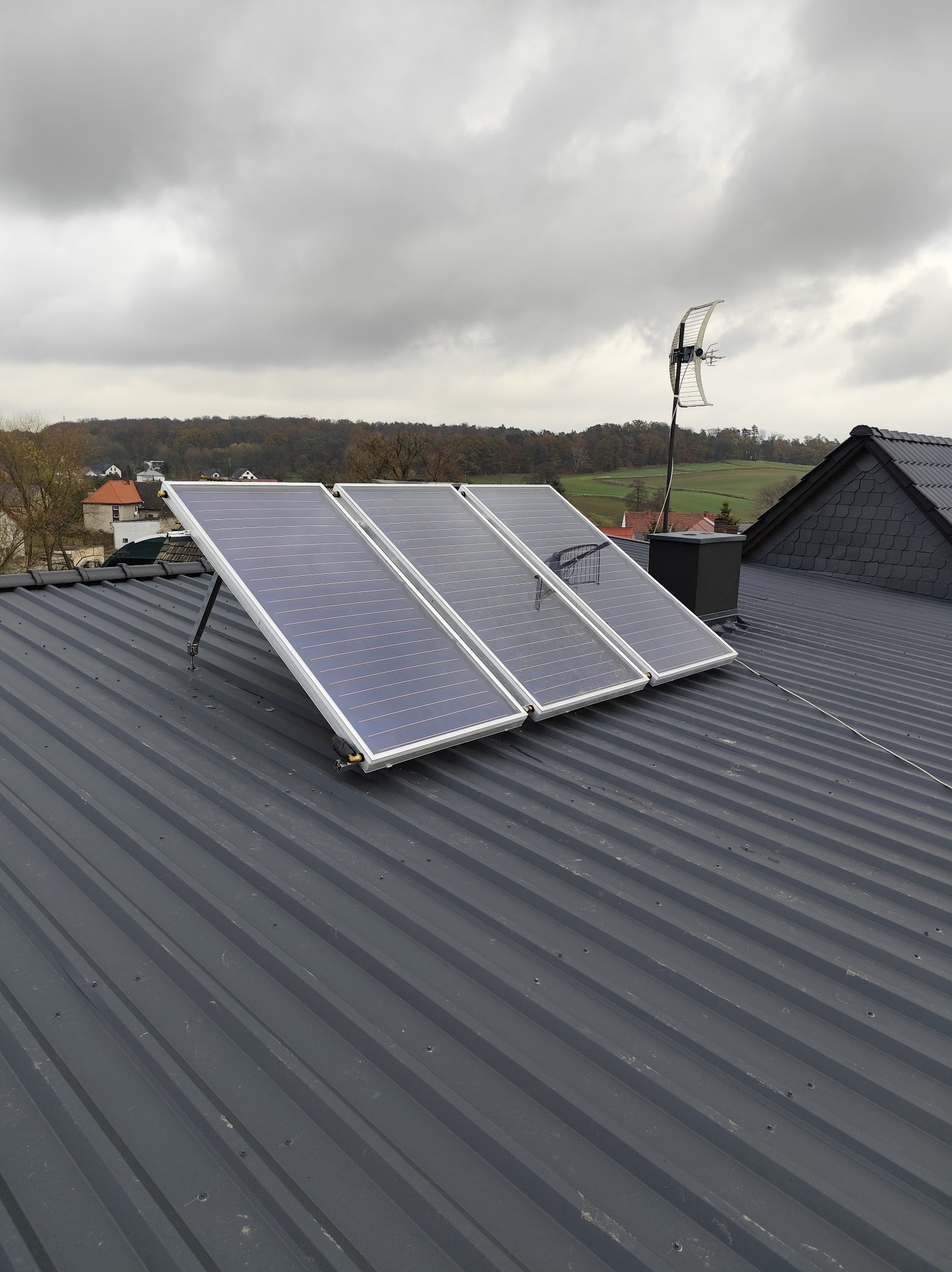 Trzy panele słoneczne zamontowane na szarym, blaszanym dachu domu z widoczną anteną i kominem na tle pochmurnego nieba i oddalonych zabudowań.