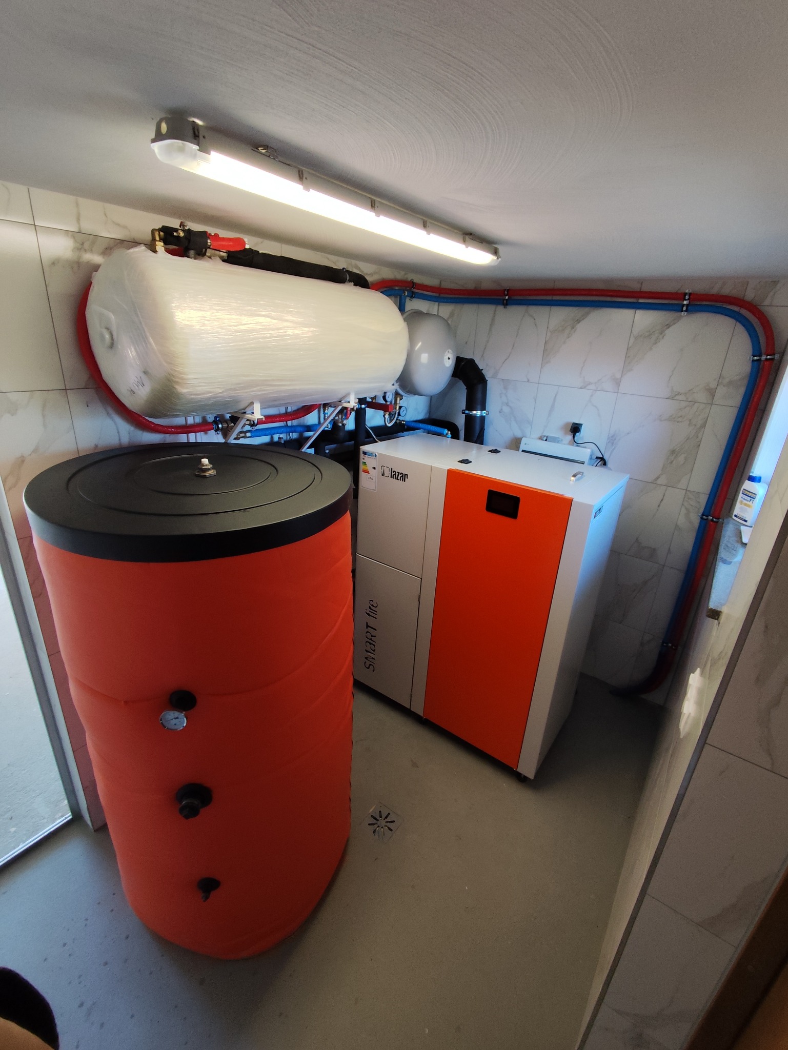 Kotłownia z piecem na pellet Lazaro Smart Fire, zasobnikiem ciepłej wody w pomarańczowej obudowie, zbiornikiem buforowym owiniętym folią i instalacją grzewczą z rurami w kolorach czerwonym...