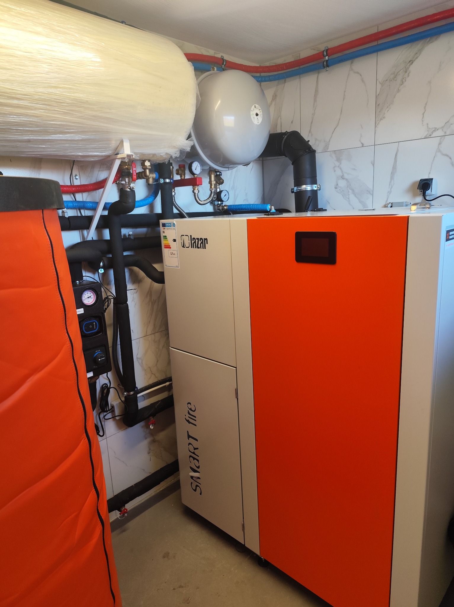 Nowoczesny piec grzewczy Lazar Smart Fire z pomarańczowym panelem frontowym i czarnym wyświetlaczem, zainstalowany w kotłowni z marmurowymi płytkami ściennymi i widocznymi rurami instalacji grzewczej.