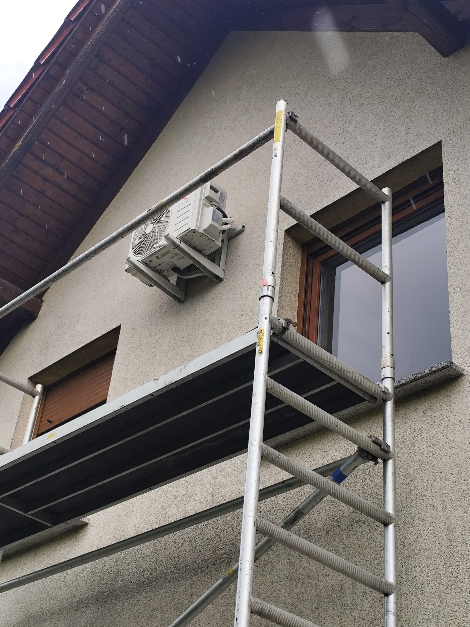 Biała jednostka zewnętrzna klimatyzacji zamontowana na elewacji domu, widoczna z rusztowania. Okno z brązowymi roletami w tle.