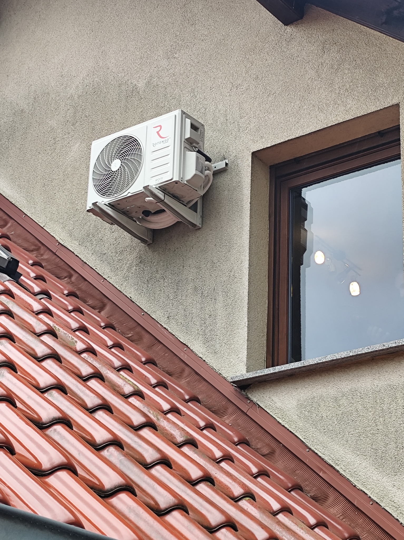 Biały klimatyzator Rotenso zamontowany na elewacji budynku, widok z dołu spod dachu. Widoczne okno z odbiciem oświetlenia w szybie.