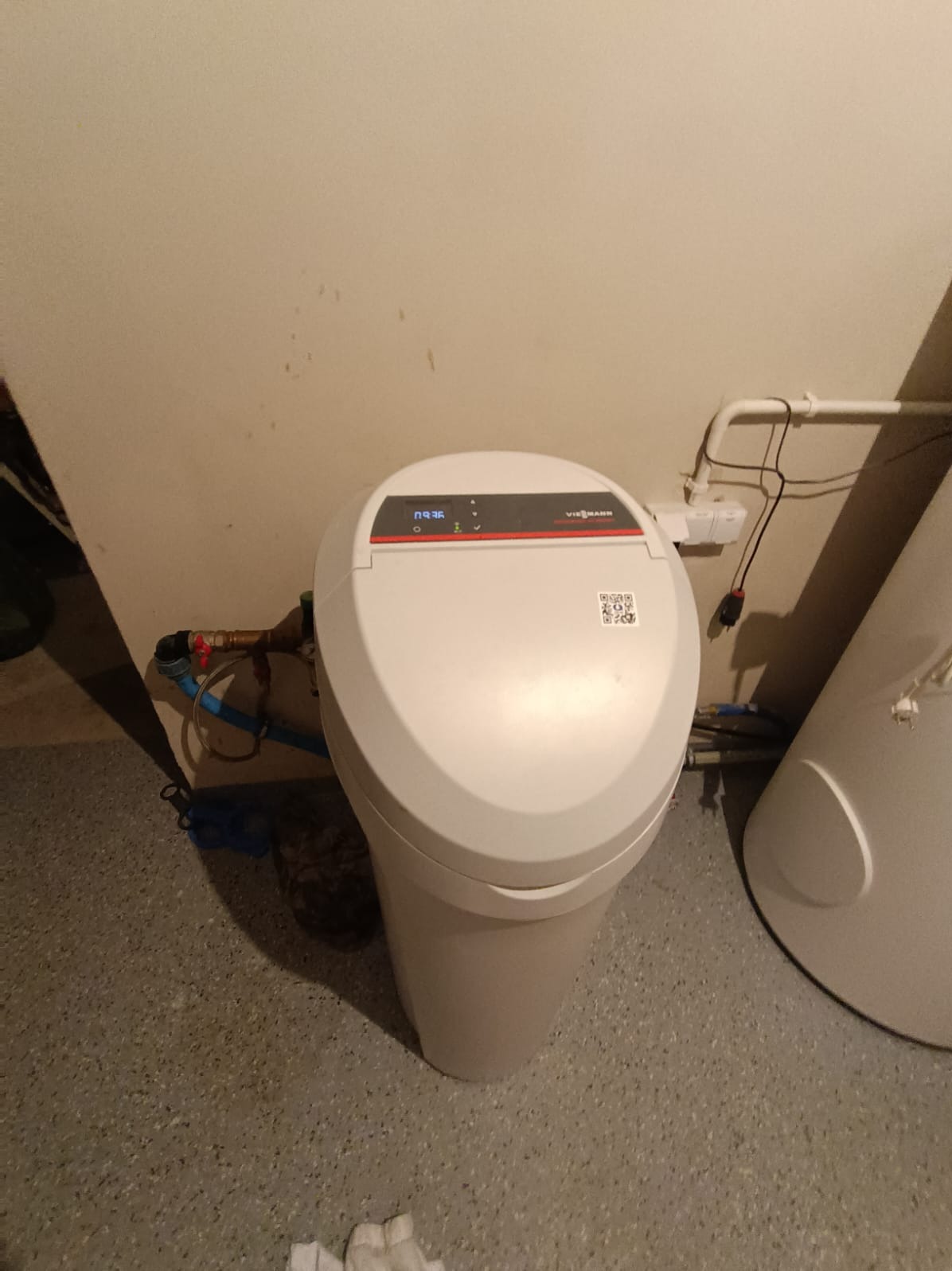 Biały zmiękczacz wody Viessmann z wyświetlaczem i kodem QR, zainstalowany przy ścianie, widoczne rury doprowadzające wodę. Obok zasobnik ciepłej wody.