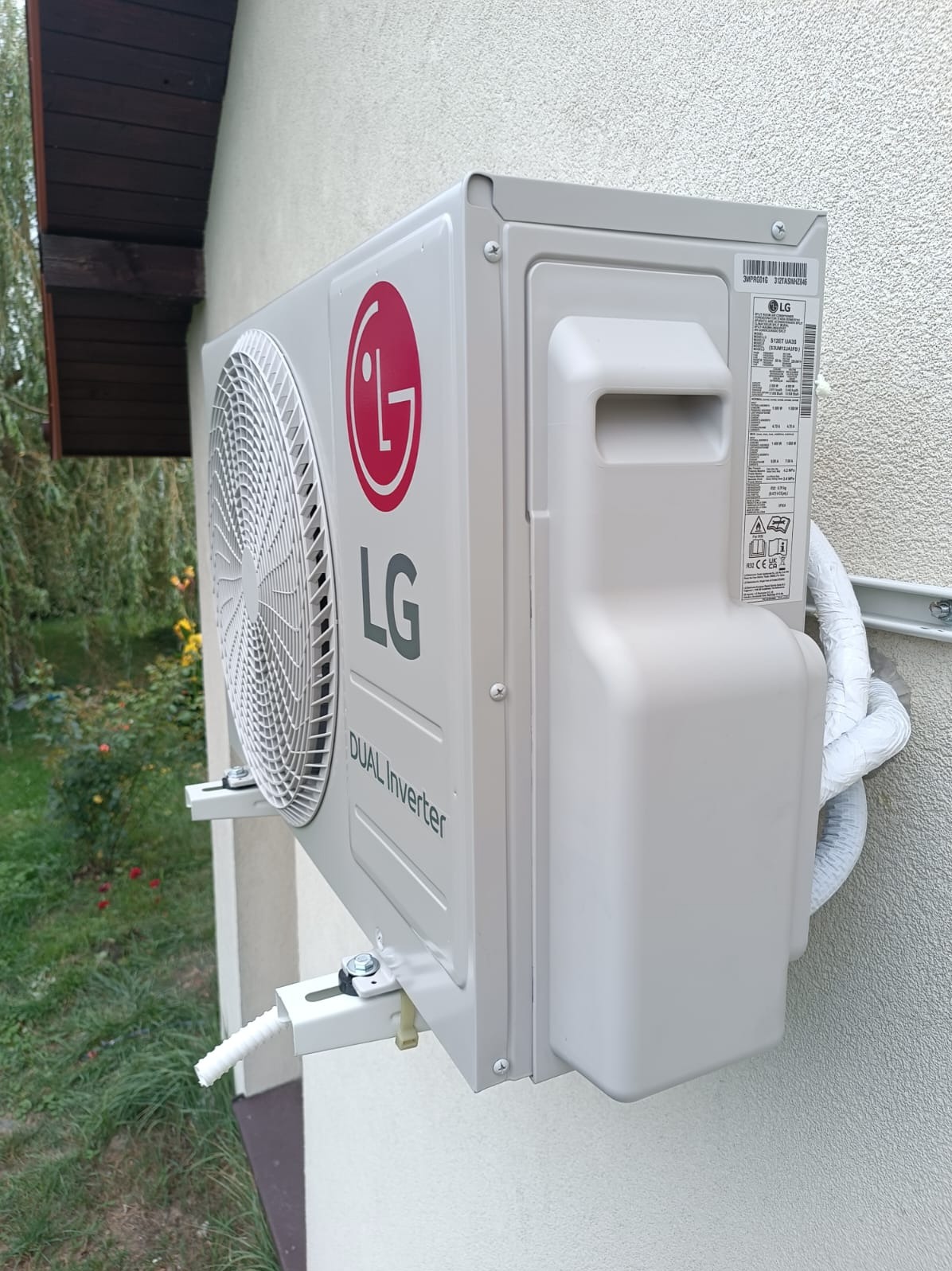 Zamontowana klimatyzacja LG Dual Inverter na elewacji budynku. Widoczne przewody w białej izolacji i elementy mocujące. Ujęcie z boku, zieleń w tle.