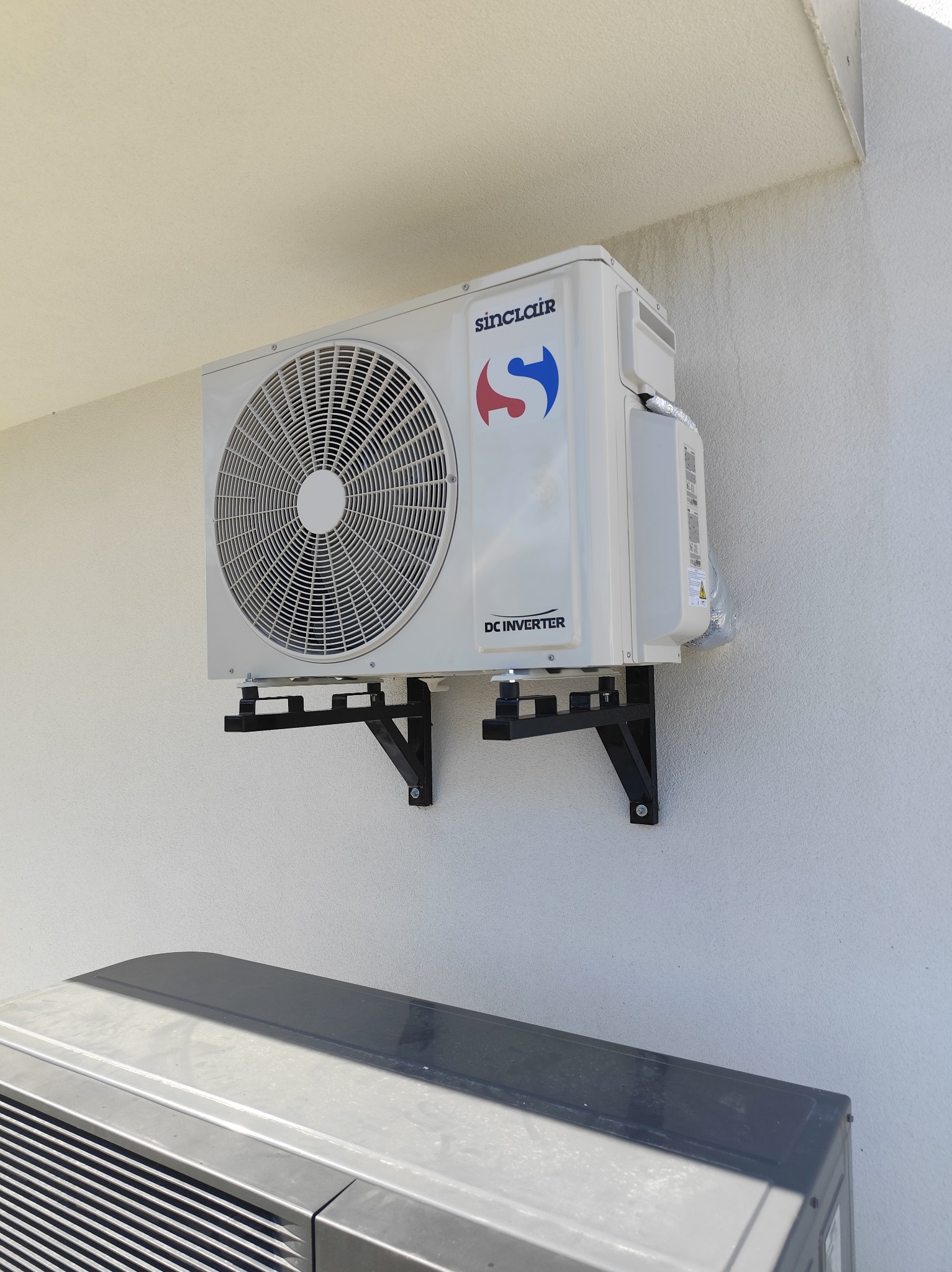 Biała jednostka zewnętrzna klimatyzacji Sinclair zamontowana na czarnych wspornikach na jasnej, teksturowanej ścianie. Widoczny fragment drugiej jednostki.