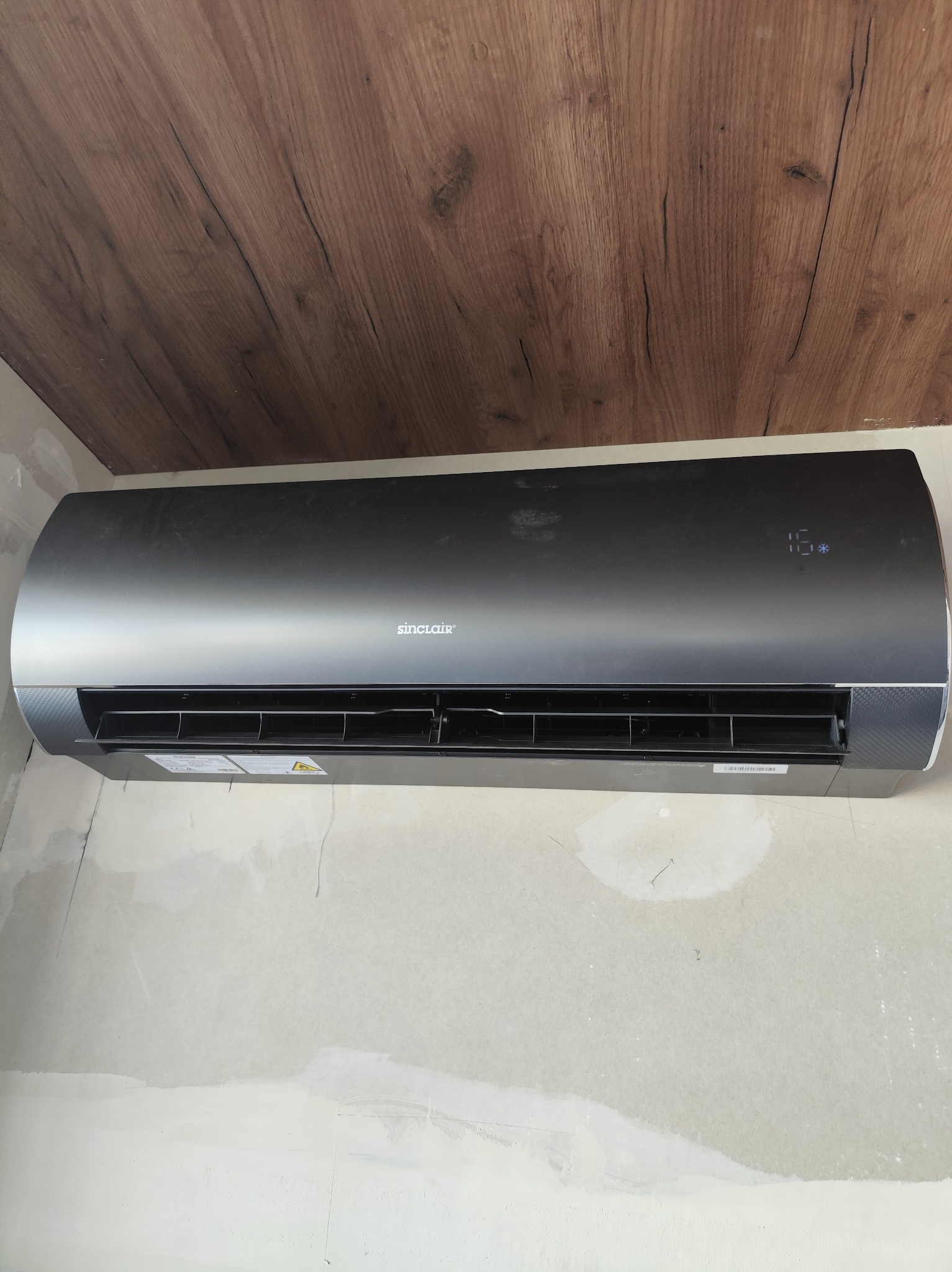 Czarny klimatyzator Sinclair na ścianie w trakcie wykończenia, widoczne drewniane panele sufitowe i niedokończone ściany. Wyświetlacz pokazuje 16 stopni.