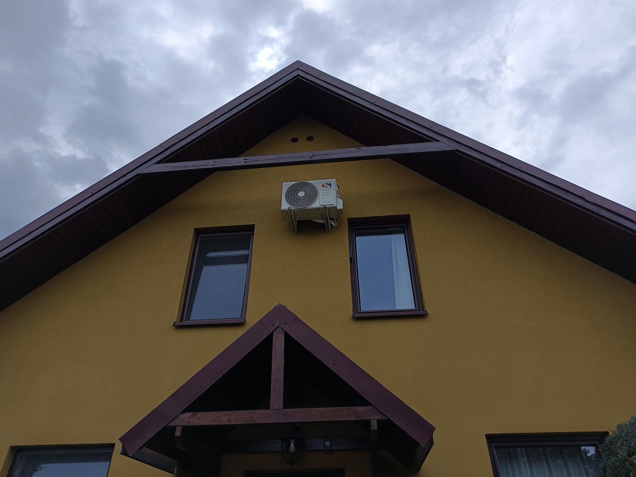 Żółta fasada domu z zamontowaną klimatyzacją zewnętrzną pod okapem dachu. Widoczne dwa okna i zadaszone wejście. Pochmurne niebo w tle.