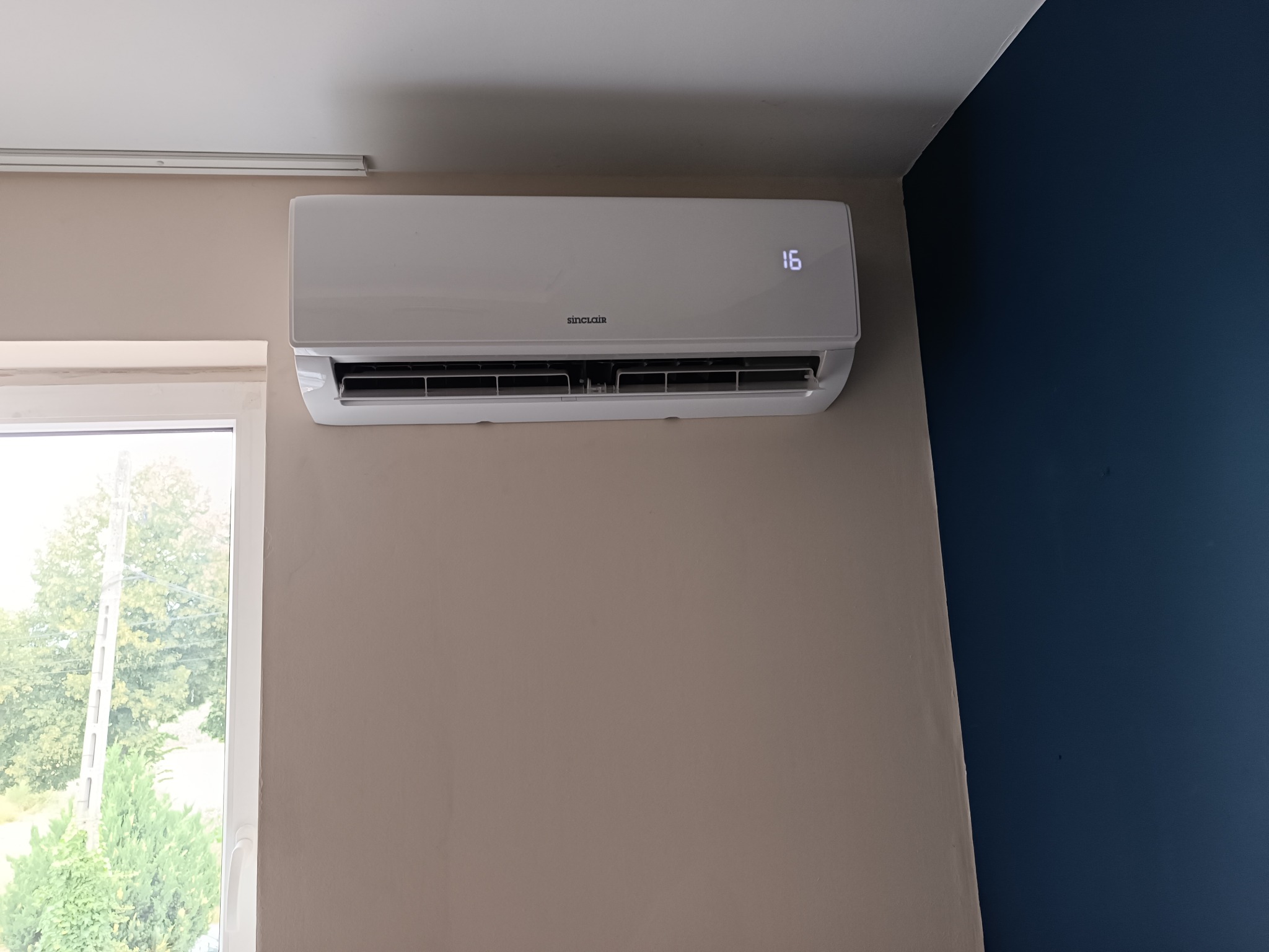 Biały klimatyzator Sinclair na beżowej ścianie, z wyświetlaczem temperatury 16 stopni i widocznym oknem z zielenią na zewnątrz. Część ściany pomalowana na granatowo.