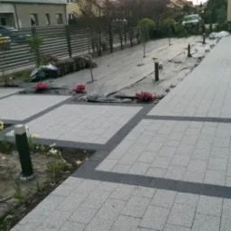 Ułożona jasnoszara kostka brukowa z ciemnoszarą obwódką tworząca kwadratowe segmenty chodnika i podjazdu, częściowo obsadzona roślinami i niskimi lampami ogrodowymi, w tle przygotowany teren...