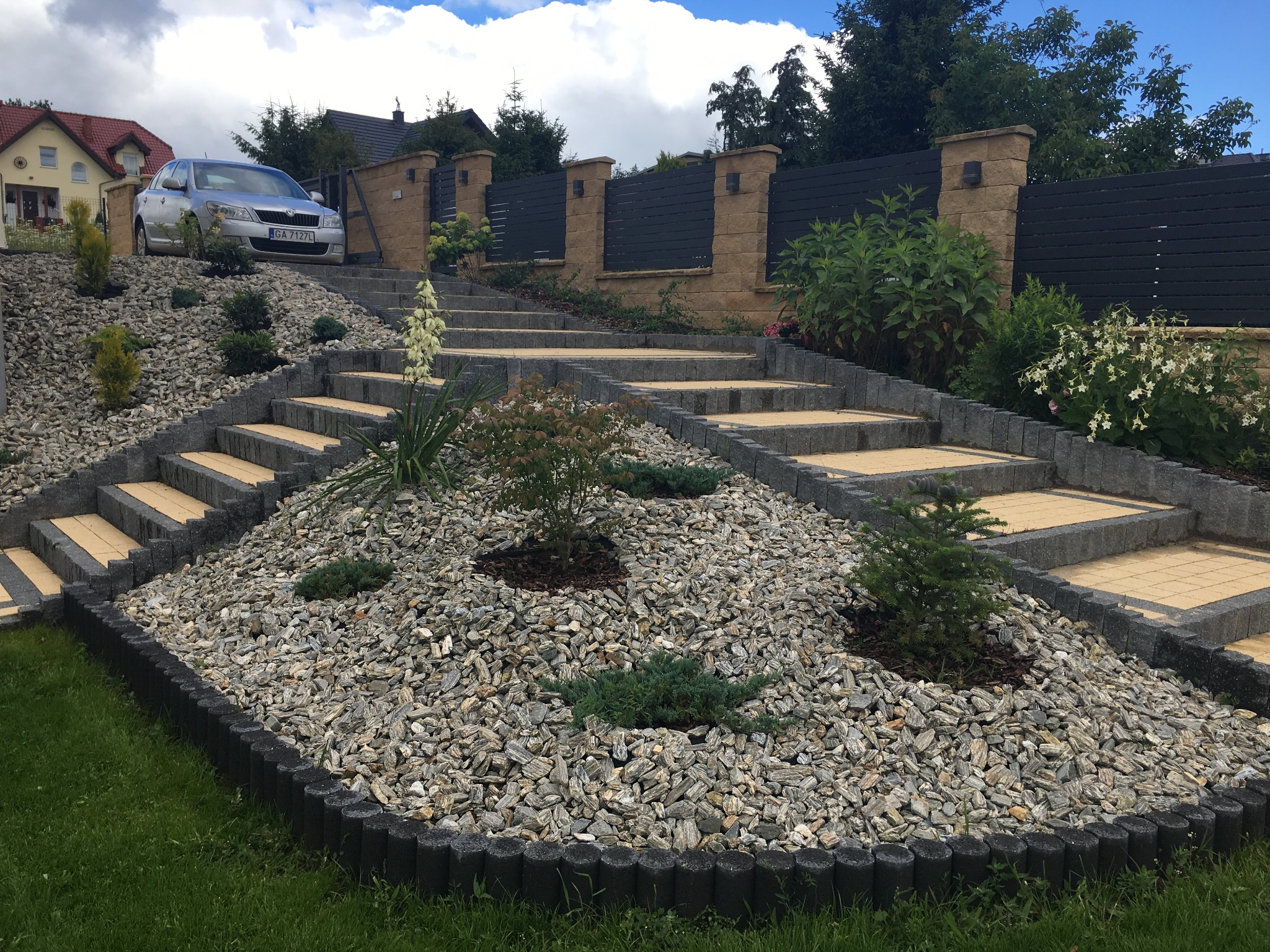 Ogród na zboczu z kamieniami ozdobnymi, schodami z kostki brukowej w kolorze piaskowym, obrzeżami z betonowych palisad i roślinami, w tle szary samochód osobowy i nowoczesne ogrodzenie z elementami...