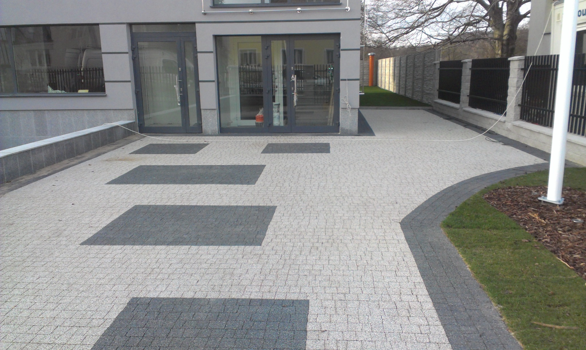 Jasnoszara kostka brukowa ułożona w geometryczny wzór z ciemnoszarymi prostokątami przed nowoczesnym budynkiem z szarą elewacją i szklanymi drzwiami.