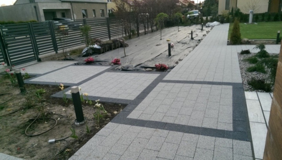 Ułożona jasnoszara kostka brukowa z ciemnoszarą obwódką tworząca kwadratowe segmenty chodnika i podjazdu, częściowo obsadzona roślinami i niskimi lampami ogrodowymi, w tle przygotowany teren...