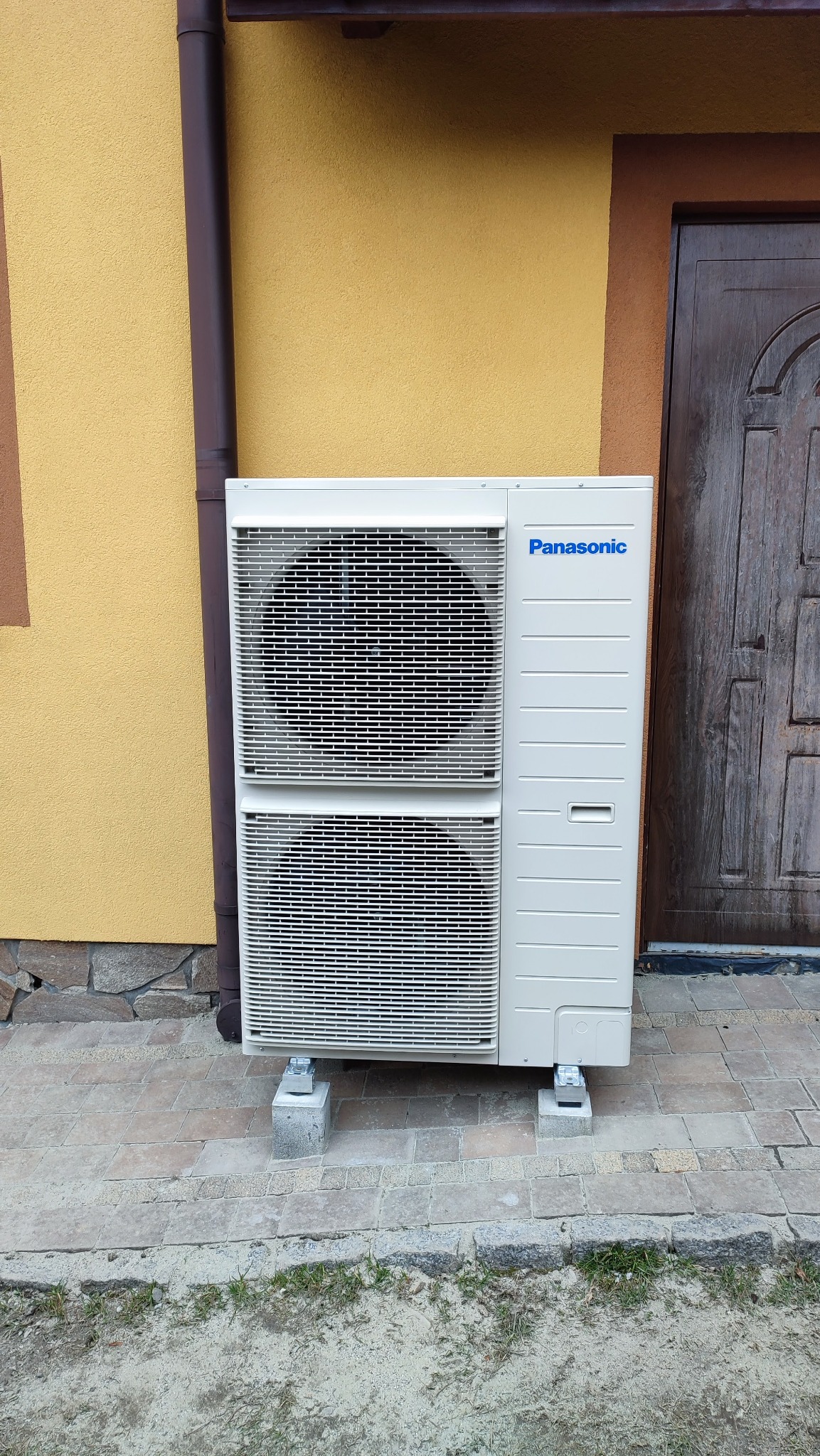 Zewnętrzna jednostka pompy ciepła Panasonic, zamontowana na betonowych bloczkach przed budynkiem z żółtą elewacją i brązowymi elementami wykończeniowymi.