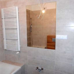 M&L Group Igor Leszczyński - Wykończona łazienka z beżowymi płytkami, grzejnikiem drabinkowym, lustrem odbijającym prysznic i drewnianą zabudowę, widoczne wystające kable elektryczne.