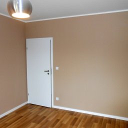 M&L Group Igor Leszczyński - Pomalowane na beżowo wnętrze pokoju z drewnianą podłogą, białą listwą przypodłogową i białą, zamkniętą ścianą z drzwiami. Na suficie widoczna metalowa lampa.