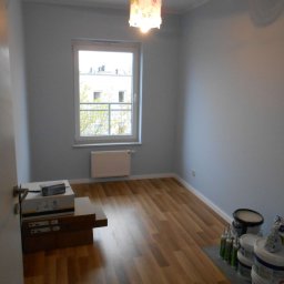 M&L Group Igor Leszczyński - Pomieszczenie po malowaniu ścian na jasny szary kolor, widoczne wiaderka z farbą, tuby z silikonem oraz podłoga z paneli.