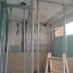 Stelaż z profili stalowych przygotowany do montażu płyt gipsowo-kartonowych w trakcie prac wykończeniowych, widoczne instalacje elektryczne i hydrauliczne, fragment drabiny malarskiej.