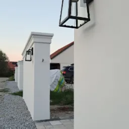 Biały mur z dekoracyjnymi słupkami i czarnymi, nowoczesnymi lampami zewnętrznymi, obok ścieżka z kostki brukowej i szarego żwiru.