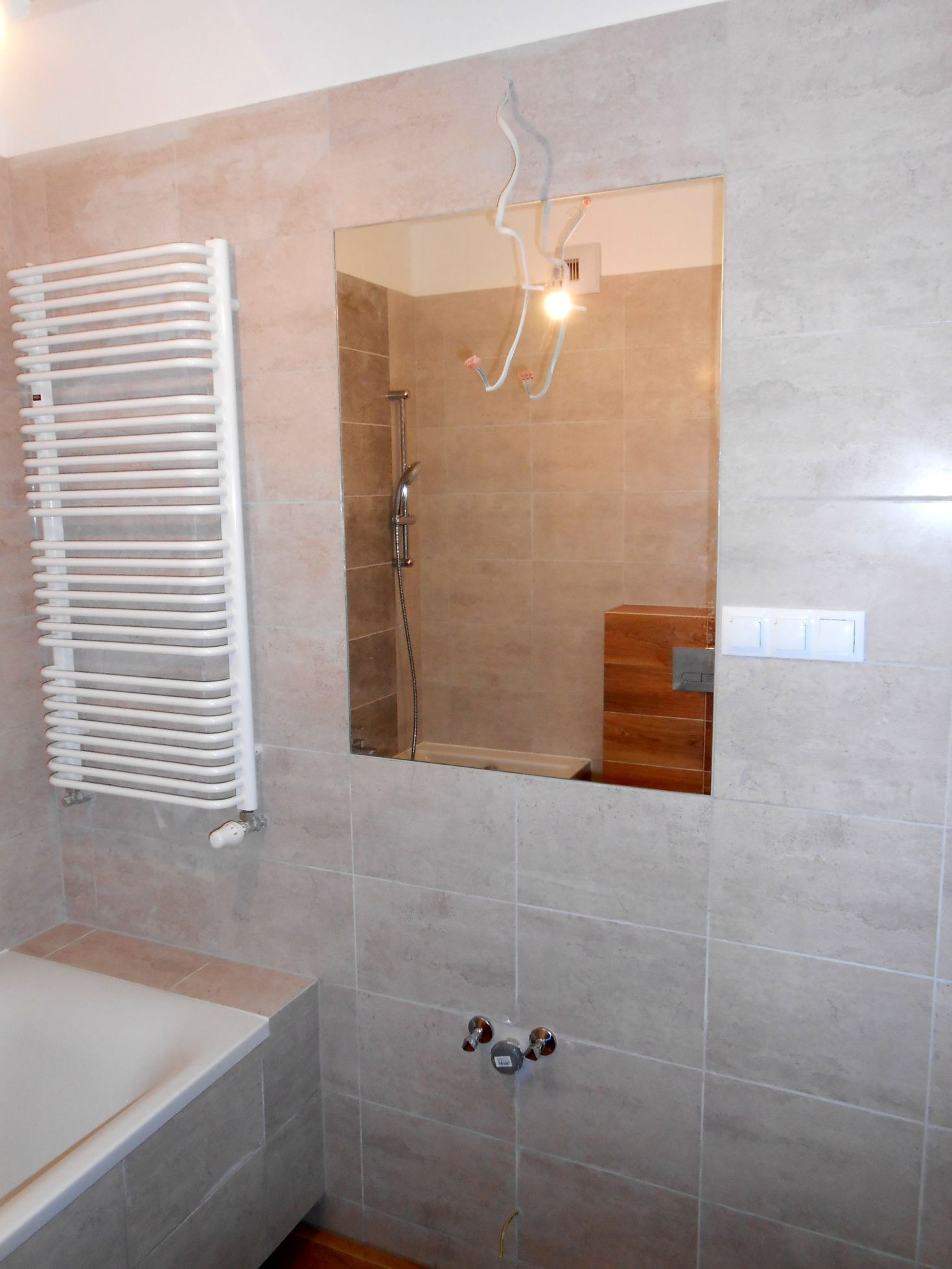 Wykończona łazienka z beżowymi płytkami, grzejnikiem drabinkowym, lustrem odbijającym prysznic i drewnianą zabudowę, widoczne wystające kable elektryczne.
