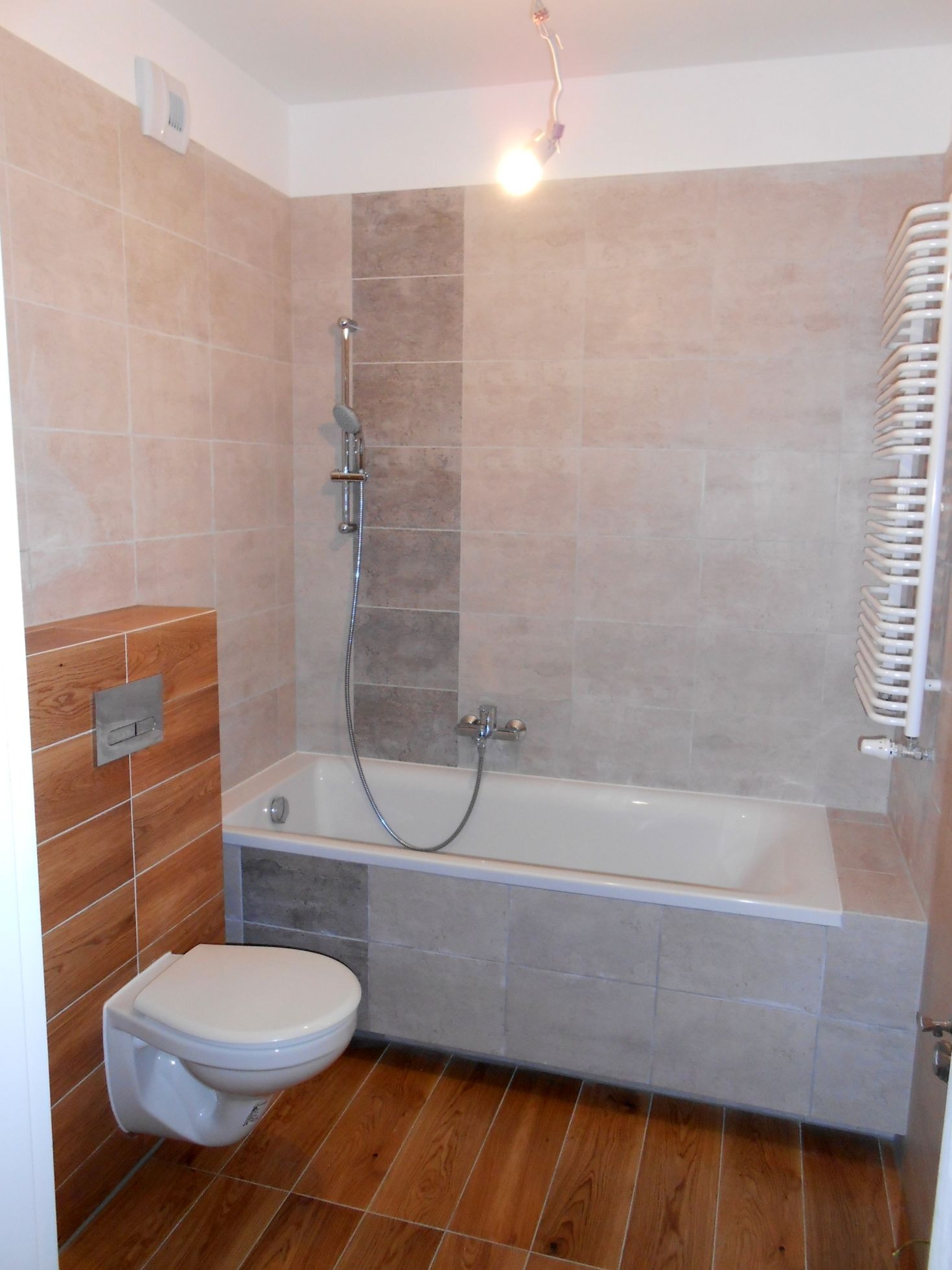 Wykończona łazienka z beżowymi i szarymi płytkami, drewnopodobną zabudową toalety i grzejnikiem drabinkowym.