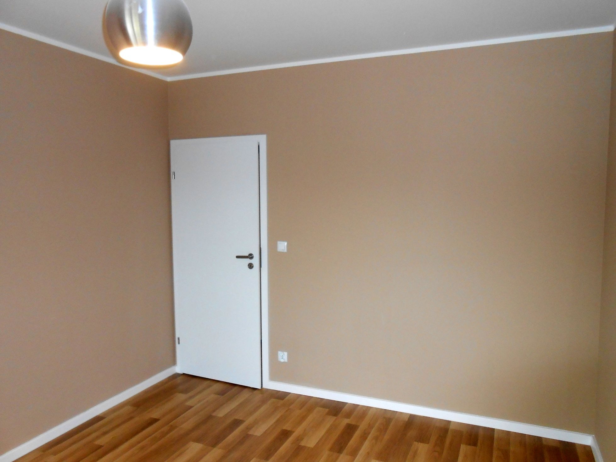 Pomalowane na beżowo wnętrze pokoju z drewnianą podłogą, białą listwą przypodłogową i białą, zamkniętą ścianą z drzwiami. Na suficie widoczna metalowa lampa.