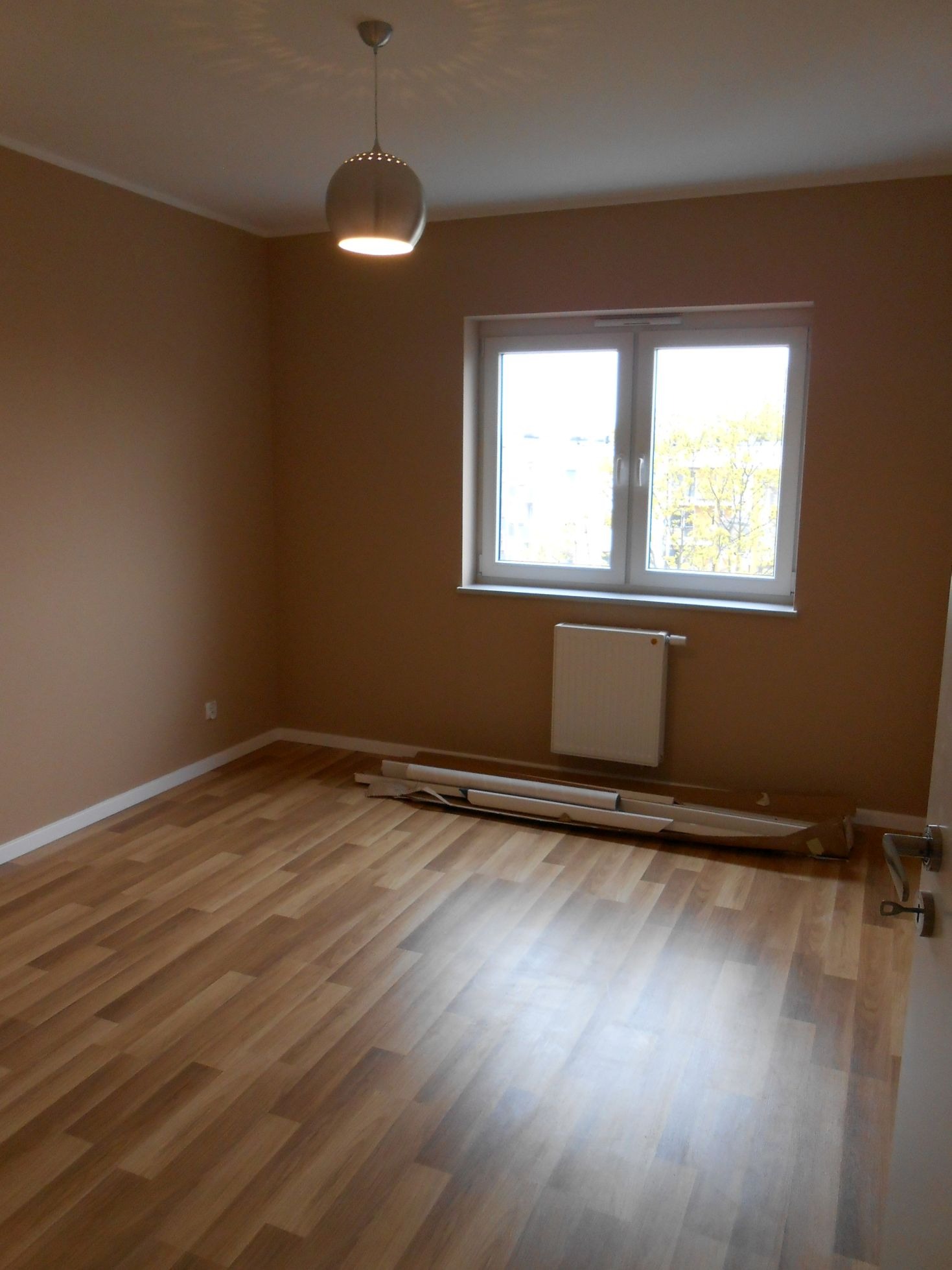 Pusty pokój z panelami podłogowymi w odcieniach brązu, beżowe ściany, białe okno z widokiem na drzewa, nowoczesna lampa sufitowa i grzejnik pod oknem. Na podłodze leżą zwinięte arkusze materiału...