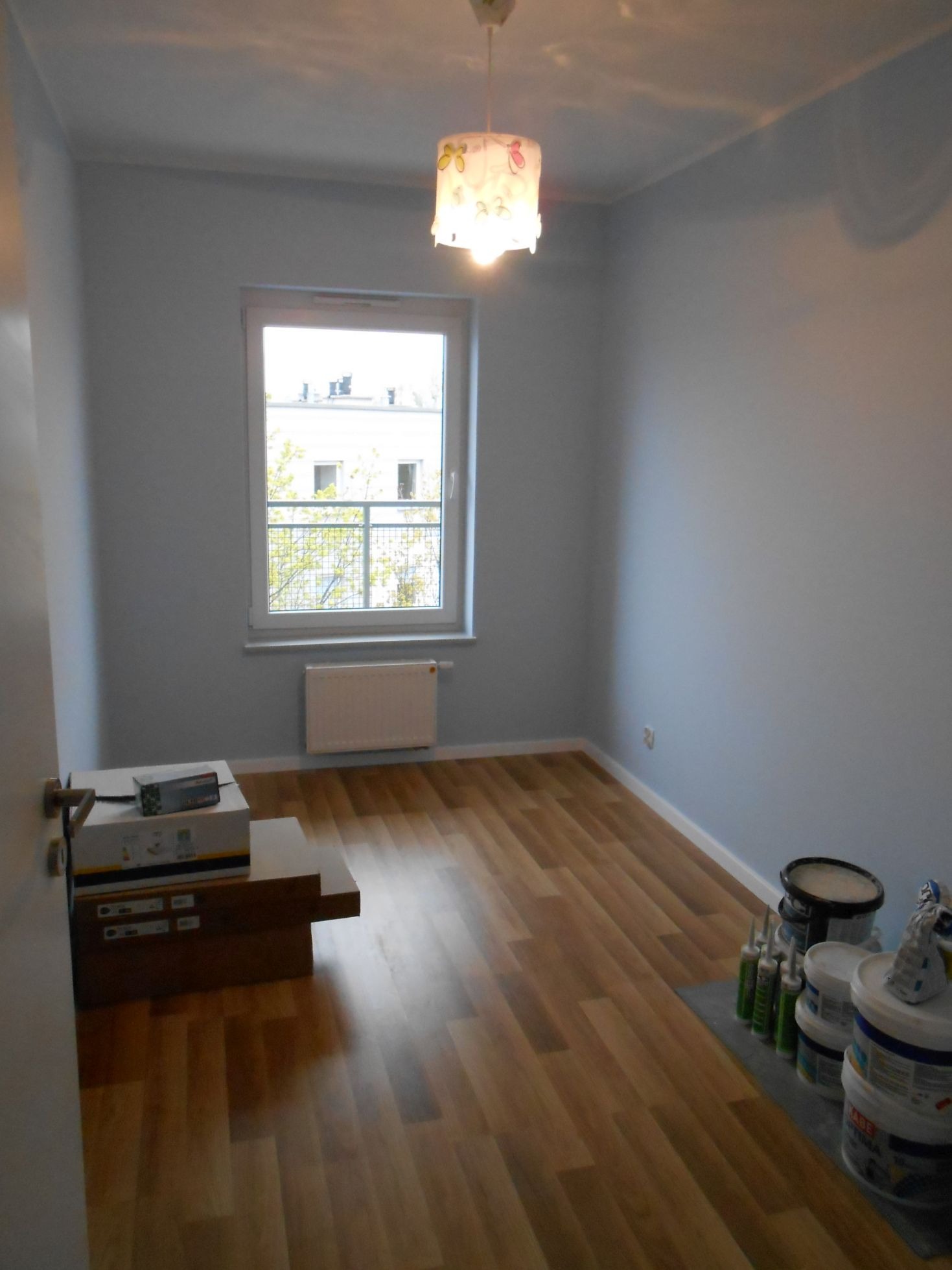 Pomieszczenie po malowaniu ścian na jasny szary kolor, widoczne wiaderka z farbą, tuby z silikonem oraz podłoga z paneli.