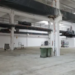 Wnętrze hali przemysłowej w trakcie odświeżania z pomalowanymi na biało ścianami i stropem, zabezpieczonymi rurami wentylacyjnymi, pomarańczowy podnośnik koszowy i zielone kontenery na śmieci w tle.