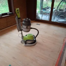 Świeżo wycyklinowany parkiet z maszyną do cyklinowania w tle, widoczny fragment szlifierki i kawałek użytego papieru ściernego na podłodze.