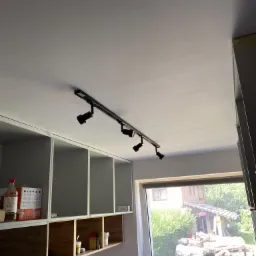 Czarna listwa z czterema reflektorami zamontowana na białym suficie w kuchni, widok na okno z widokiem na ogród i stertę drewna.