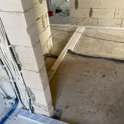 Wnętrze w trakcie remontu: surowa ściana z betonu komórkowego, wiązka kabli elektrycznych prowadzona wzdłuż ściany, listwy przypodłogowe z tworzywa sztucznego leżące na podłodze, widoczne narzędzia...