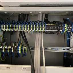 Wnętrze szarej szafy elektrycznej z widocznymi listwami zaciskowymi, do których podłączone są przewody w kolorach czarnym, niebieskim, brązowym i żółto-zielonym. Przewody ułożone w uporządkowany...