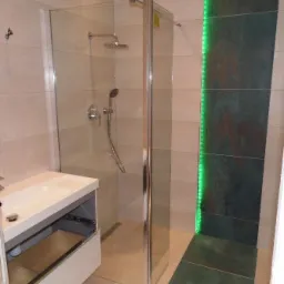 Nowoczesna łazienka z kabiną prysznicową typu walk-in, umywalką z szafką z otwartą szufladą i zielonym oświetleniem LED na ścianie z ciemnych płytek.