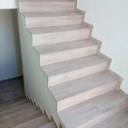 Schody wewnętrzne wyłożone płytkami imitującymi drewno, z charakterystycznym wykończeniem krawędzi stopni i przypodłogowym, widok z dołu.