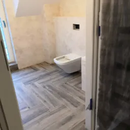 Niedokończona łazienka z ułożoną szarą podłogą w jodełkę, wisząca toaleta i przygotowane ściany pod płytki, widok przez uchylone drzwi zabezpieczone folią.