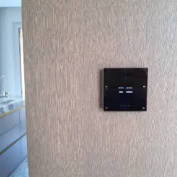 Czarny panel dotykowy systemu inteligentnego domu marki Grenton, zamontowany na ścianie z drewnianym wykończeniem, z widocznym fragmentem blatu kuchennego w tle.