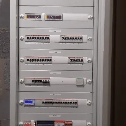 Otwarta szafa elektryczna z panelami sterowania, wyłącznikami, zabezpieczeniami przepięciowymi, licznikiem energii i akumulatorem podtrzymującym zasilanie awaryjne.