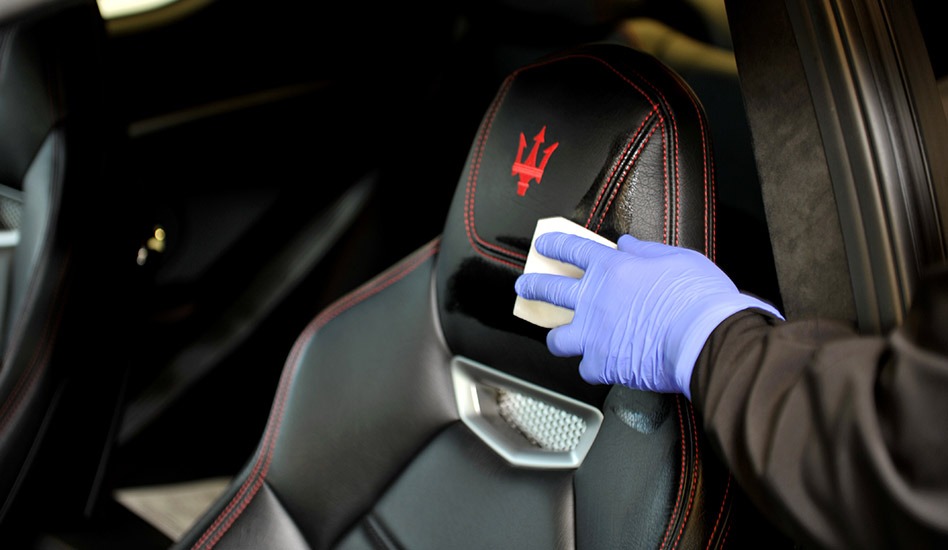 Czyszczenie skórzanego fotela samochodowego z czerwonym logo trójzębu, widoczna ręka w niebieskiej rękawiczce aplikująca preparat.