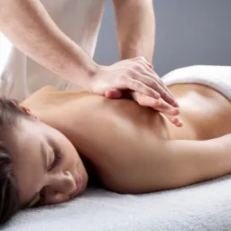 Kobieta leży na brzuchu na białym ręczniku, a terapeuta w białym stroju wykonuje masaż górnej części pleców, używając obu rąk.