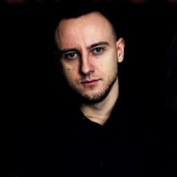 Portret mężczyzny z krótką brodą i ciemną koszulą na czarnym tle, z fragmentem kobiecej sylwetki po prawej stronie kadru.