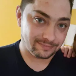 Portret mężczyzny z krótką, ciemną brodą i niebieskimi oczami, ubrany w czarną koszulkę na tle żółtej ściany.