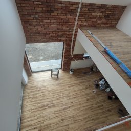 Złota Rączka - Wnętrze domu w trakcie prac wykończeniowych: ściana z cegły klinkierowej, drewnopodobna podłoga, okno dachowe, widoczne narzędzia i puszki po farbach, niebieska folia ochronna na schodach.