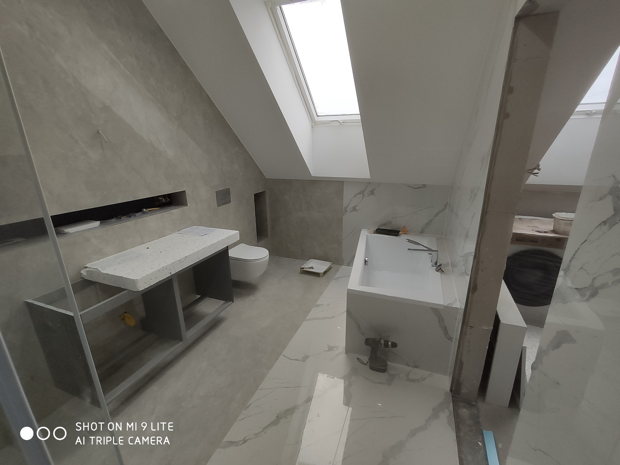 Nowoczesna łazienka na poddaszu ze skośnym oknem dachowym, wykończona płytkami imitującymi marmur, z białą wanną, toaletą w zabudowie i umywalką na szarym stelażu. Na podłodze leży wkrętarka.
