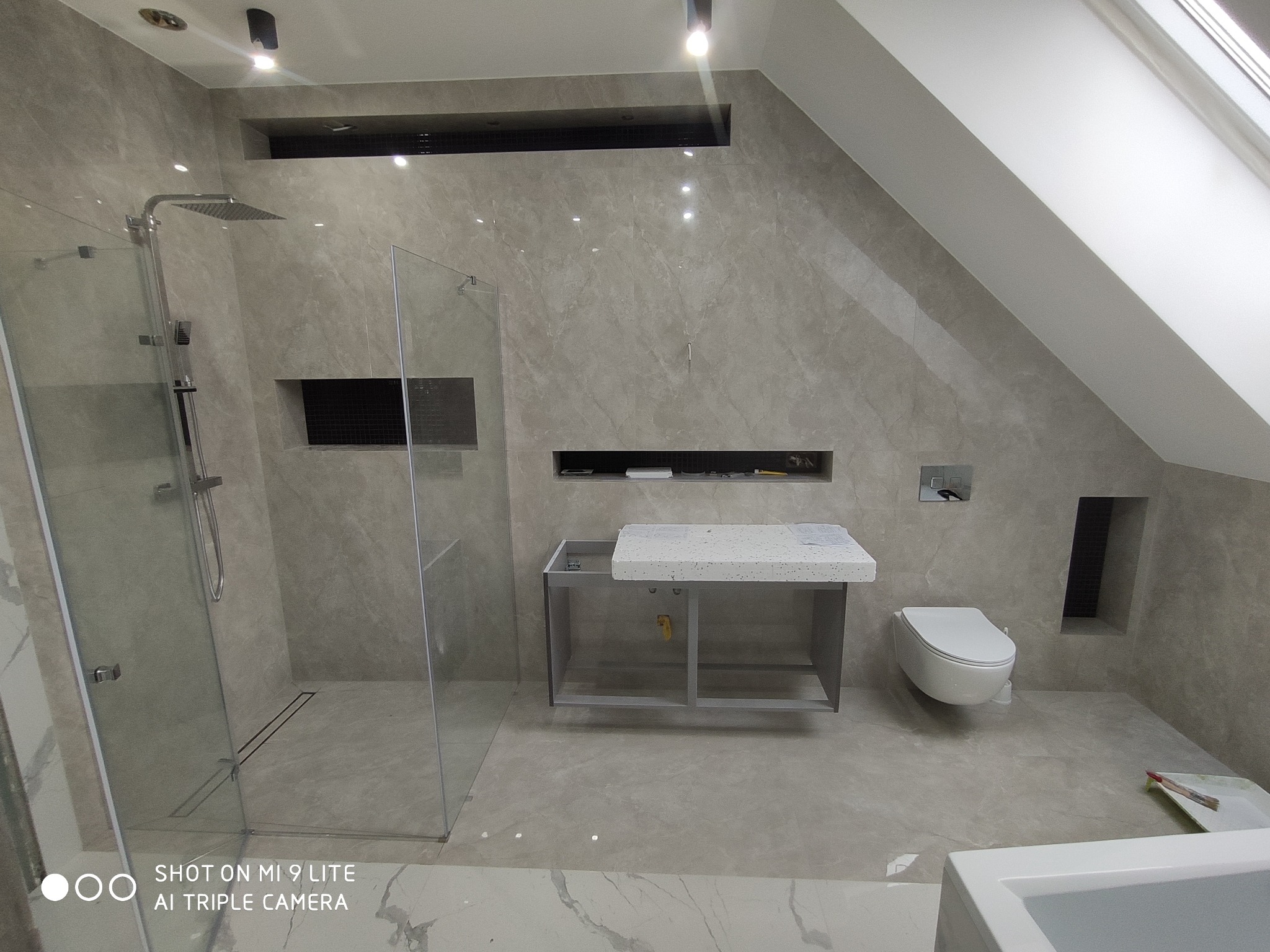 Nowoczesna łazienka na poddaszu w odcieniach szarości z kabiną prysznicową typu walk-in, wiszącą toaletą i umywalką na metalowym stelażu z blatem z lastryko.